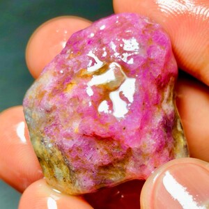 以下が含まれることがあります： 鮮やかなマゼンタとピンクの色合いを持つ、未研磨の原石。自然な鉱物の内包物と土のような茶色の斑点がある質感の不均一な表面で、不規則な有機的形状全体に濡れたような光沢のある輝きを放っています。