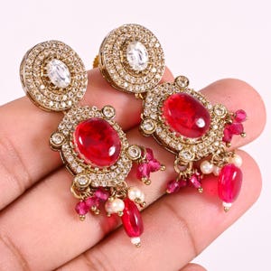 May include: Gold-toned earrings with a central oval red gemstone, surrounded by small clear stones. The earrings have a circular top and dangling red and white accents. The earrings are held in a hand.