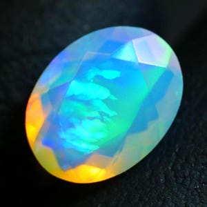 Può includere: Una gemma di opale di forma ovale con taglio sfaccettato. La gemma mostra un gioco di colori, con lampi di blu, verde, giallo e arancione. La pietra è posta su uno sfondo scuro, evidenziando le sue qualità iridescenti.