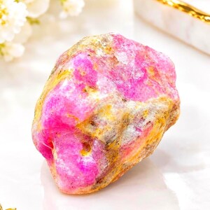 Peut inclure: Un spécimen minéral brut et non poli présentant des taches vibrantes de rose vif entremêlées de dépôts minéraux terreux jaunes, fauves et gris. La pierre a une forme organique irrégulière avec une surface texturée et inégale.