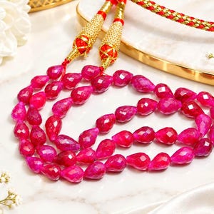 Könnte beinhalten: Eine mehrsträngige Halskette mit facettierten, tropfenförmigen fuchsiafarbenen Perlen. Die Kette verfügt über zwei dekorative, goldfarbene Metallendkappen mit komplexen Texturen und roten Akzenten, die an einem geflochtenen rot-goldenen Kordelverschluss befestigt sind.