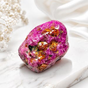 Könnte beinhalten: Ein rohes, unregelmäßig geformtes Mineralexemplar mit einer leuchtend magentafarbenen Grundfarbe, eingebetteten, durchscheinenden, bernsteinfarbenen kristallinen Einschlüssen und kleinen dunklen Mineralflecken auf der texturierten, polierten Oberfläche.
