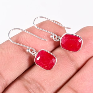 Op de afbeelding: Een paar zilveren oorbellen met helderrode, rechthoekige edelstenen. De edelstenen zijn facetgeslepen en vangen het licht. De oorbellen worden gepresenteerd tegen een neutrale achtergrond, waardoor de levendige kleur van de stenen wordt benadrukt.