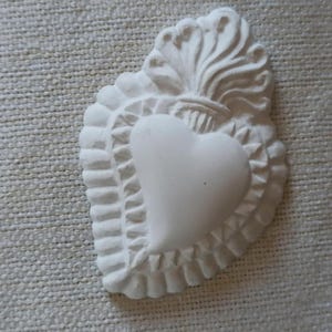 Può includere: Un elemento decorativo bianco a forma di cuore con una superficie strutturata. Il design presenta un cuore centrale, circondato da un bordo smerlato e sormontato da una fiamma stilizzata o un elemento floreale. Il pezzo è probabilmente realizzato in gesso o materiale simile.