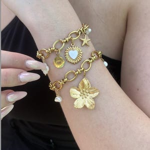 Peut inclure: Bracelets à breloques dorés avec divers pendentifs, dont un tournesol, une étoile, une fleur et un cœur. Les bracelets comportent également de petites perles pendantes. Les bijoux sont portés au poignet.
