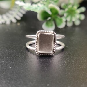May include: A silver ring featuring a rectangular bezel setting with a twisted rope-style border. The ring has a split shank design, consisting of two thin, smooth bands that connect at the sides to support the central rectangular frame.