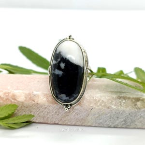 May include: A silver ring featuring a large, oval-shaped stone with a striking black and white pattern. The stone is set in a detailed silver band, creating a bold and elegant statement piece. The ring is displayed on a light-colored surface.