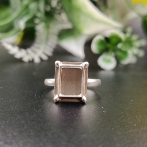 May include: A silver-toned ring featuring a rectangular setting with four prongs at the corners. The center of the setting is flat and recessed, designed to hold a stone. The band is smooth and metallic, connecting to the base of the rectangular frame.