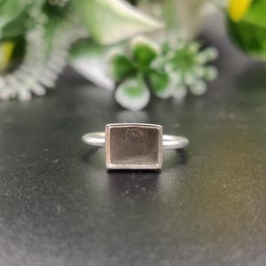 May include: A silver-toned ring featuring a rectangular bezel setting with a flat, recessed center. The band is thin and smooth, connecting to the sides of the rectangular frame. The metal has a polished finish, creating a clean and simple design.