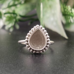 May include: A silver ring featuring a teardrop-shaped setting with a smooth, matte gray center inlay. The bezel is surrounded by a decorative border of small, raised silver spheres, and the band is a simple, polished silver wire design.
