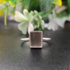 May include: A silver-toned ring featuring a rectangular bezel setting with a flat, dark gray, matte-finished inlay. The band is thin and smooth, connecting to the sides of the rectangular frame, which has slightly rounded corners and a polished edge.