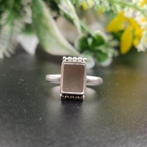 May include: A silver ring featuring a rectangular setting with a smooth, flat, matte-finish center panel. The top and bottom edges of the rectangular frame are decorated with a row of small, raised silver beads, attached to a plain silver band.