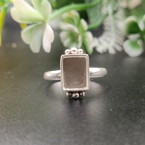 May include: A silver ring featuring a rectangular setting with a flat, dark gray inlay. The setting is accented with three small silver spheres at both the top and bottom edges. The band is smooth, polished silver, creating a clean and simple design.