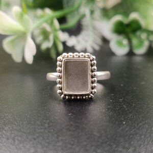 May include: A silver ring featuring a rectangular bezel setting with a smooth, flat center surface. The bezel is framed by a decorative border of small, raised silver spheres, creating a textured edge around the central rectangular area.