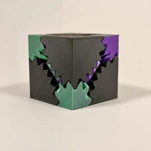 May include: A three-dimensional cube puzzle featuring a matte black exterior shell with jagged, interlocking edges. The internal components are visible through the gaps, revealing bright green and purple plastic segments with a textured finish.