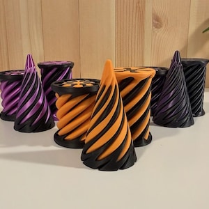 May include: A collection of 3D printed objects featuring spiral designs in black and purple or black and orange. The set includes several cylindrical spools with textured tops and pointed conical shapes, all arranged together on a flat surface.