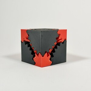 May include: A geometric puzzle cube constructed from interlocking matte black and vibrant red plastic components. The design features jagged, saw-toothed edges where the two colors meet, creating a complex, multi-layered structural appearance.