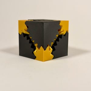 May include: A geometric puzzle cube featuring interlocking black and yellow components. The design consists of solid black corner pieces with jagged, tooth-like edges that fit into corresponding yellow sections, creating a complex, multi-layered structure.