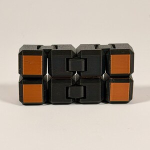 May include: A black fidget toy constructed from eight interconnected cubic segments. Each segment features a smooth, flat, square-shaped copper-colored inlay on the front face. The toy is arranged in a rectangular block formation with visible hinges.