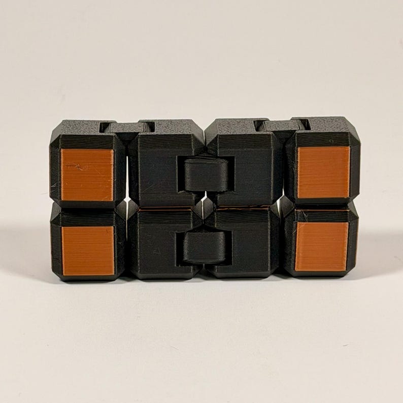 May include: A black fidget toy constructed from eight interconnected cubic segments. Each segment features a smooth, flat, square-shaped copper-colored inlay on the front face. The toy is arranged in a rectangular block formation with visible hinges.