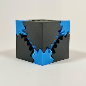 May include: A geometric puzzle cube featuring a matte black exterior shell with interlocking jagged edges that reveal a metallic blue inner core. The design consists of sharp, angular protrusions and recessed sections that fit together precisely.