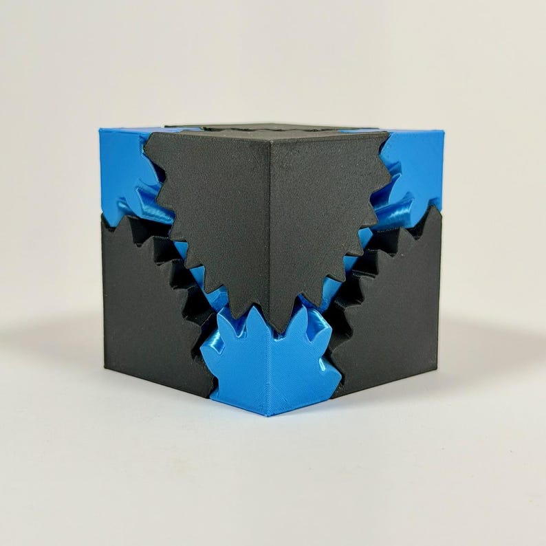 May include: A geometric puzzle cube featuring a matte black exterior shell with interlocking jagged edges that reveal a metallic blue inner core. The design consists of sharp, angular protrusions and recessed sections that fit together precisely.