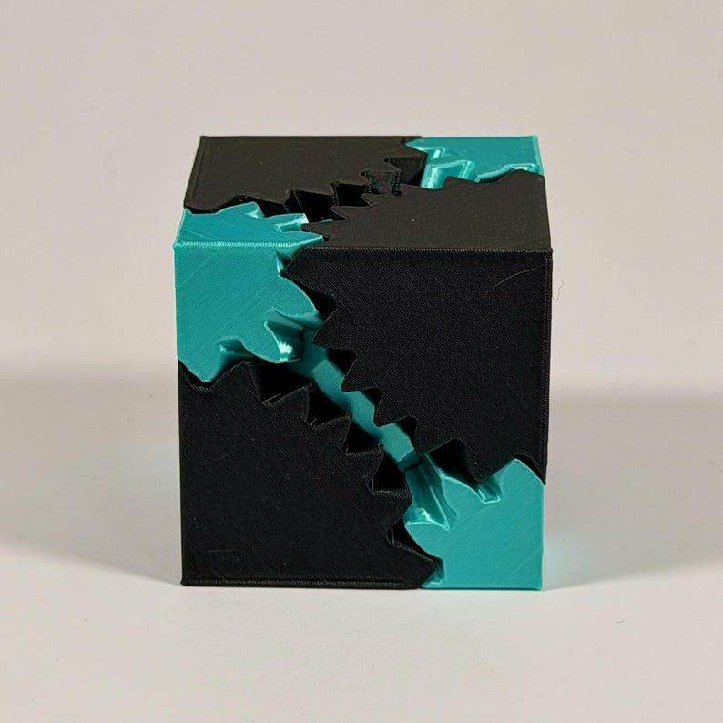 May include: A three-dimensional cube puzzle featuring interlocking geometric components. The object is constructed from matte black and glossy teal plastic segments that fit together with jagged, gear-like edges along the diagonal seams.