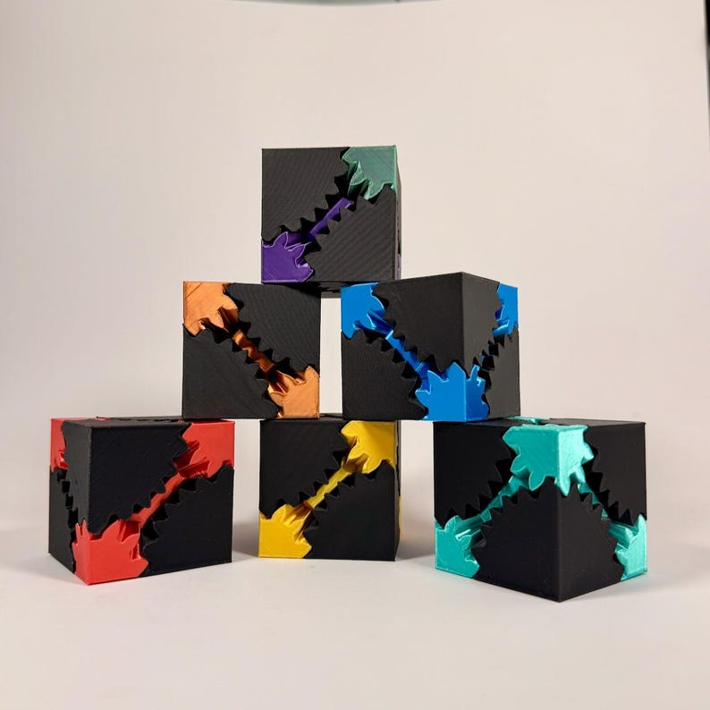May include: A stack of six black cubes featuring interlocking gear mechanisms in various colors including red, orange, yellow, green, blue, and purple. Each cube has a matte black exterior with a visible diagonal gear pattern cut out.