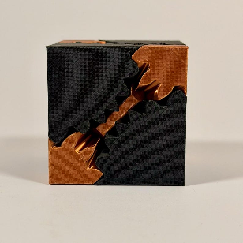 May include: A 3D printed cube puzzle featuring a two-tone design with matte black and metallic copper-colored plastic components. The cube is constructed from interlocking geometric pieces that form a diagonal jagged seam across the front face.