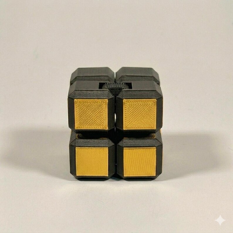 May include: A black and gold fidget cube puzzle toy featuring a modular design with four distinct square sections. The exterior frame is matte black, while each face displays a metallic gold square insert with a visible textured crosshatch pattern.