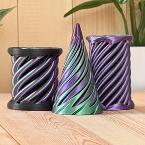 May include: Three decorative objects with a spiral design. On the left, a black cylindrical container with purple accents. In the center, a conical sculpture with a metallic purple and green finish. On the right, a purple cylindrical container.
