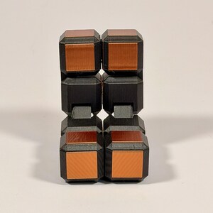 May include: A fidget toy constructed from eight interconnected cubes. Each cube features a matte black frame with a metallic copper-colored square inset on the front and top faces. The cubes are arranged in a vertical, folded rectangular configuration.