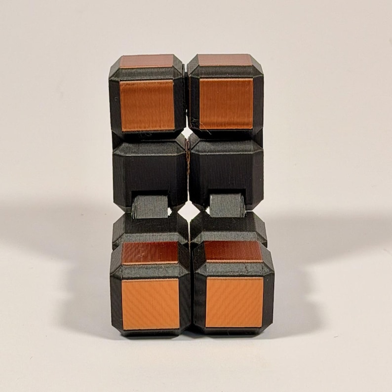 May include: A fidget toy constructed from eight interconnected cubes. Each cube features a matte black frame with a metallic copper-colored square inset on the front and top faces. The cubes are arranged in a vertical, folded rectangular configuration.