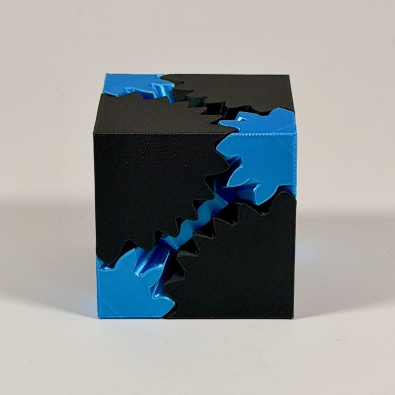 May include: A 3D printed cube puzzle featuring interlocking black and bright blue gear-shaped components. The geometric object displays a complex arrangement of jagged, teeth-like edges where the two contrasting colors meet and mesh together.