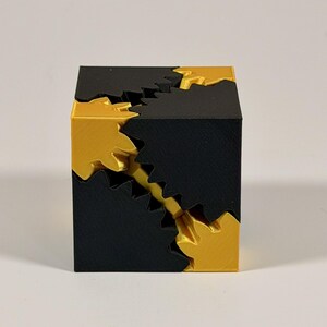 May include: A geometric cube puzzle constructed from interlocking black and gold components. The object features a series of jagged, gear-like teeth that mesh together along the diagonal edges, creating a complex, multi-part structure.