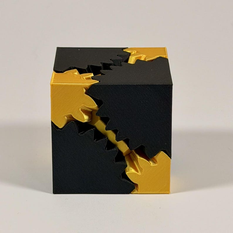 May include: A geometric cube puzzle constructed from interlocking black and gold components. The object features a series of jagged, gear-like teeth that mesh together along the diagonal edges, creating a complex, multi-part structure.