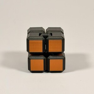 May include: A black and orange fidget cube puzzle toy featuring a modular design with four visible square faces on the front. The object is constructed from matte black plastic components with smooth, flat orange square inserts on each section.