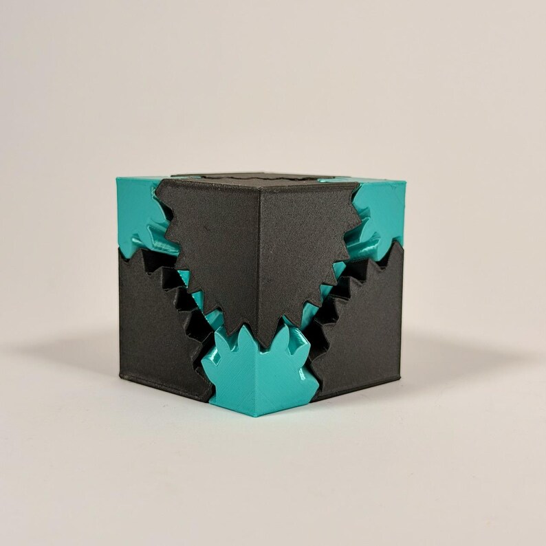 May include: A geometric cube puzzle featuring interlocking black and teal components. The design consists of solid black corner pieces that fit together with jagged, gear-like teal sections, creating a complex, multi-layered cubic structure.