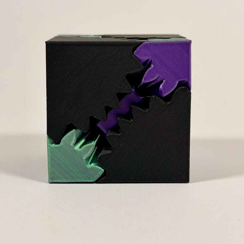 May include: A three-dimensional cube puzzle featuring a black exterior shell with a jagged, diagonal cut revealing internal components in vibrant purple and metallic green. The surface displays a textured, layered finish from 3D printing.