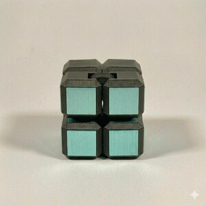 May include: A black fidget cube puzzle with a matte finish and metallic light blue square inlays on each face. The cube features a textured surface and a small black gear wheel visible between the top two segments of the puzzle structure.