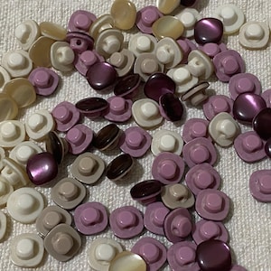 Puede incluir: Una colección de botones variados en tonos de blanco, beige, gris y morado. Los botones tienen una parte superior abovedada o plana con un vástago en la parte posterior. Algunos botones tienen un brillo nacarado. Los botones están esparcidos sobre una superficie blanca texturizada.