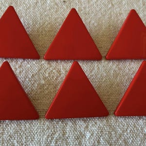 May include: Six bright red triangle-shaped game pieces arranged on a textured, off-white surface. The triangles have a glossy finish and are evenly spaced, creating a geometric pattern. These pieces could be used for a board game or educational activity.