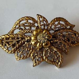 Puede incluir: Un broche de metal dorado con forma de flor, con intrincados patrones de hojas en filigrana y un único estrás redondo transparente y facetado engastado en el centro. El metal tiene un acabado mate texturizado con venas detalladas.