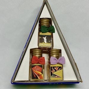 Pode incluir: Uma caixa triangular contém três pequenos frascos de perfume com tampas douradas e laços coloridos. Os frascos têm rótulos com texto e ilustrações. O frasco superior tem um laço verde, o do meio um laço vermelho e o inferior um laço roxo. A caixa é branca com borda azul.