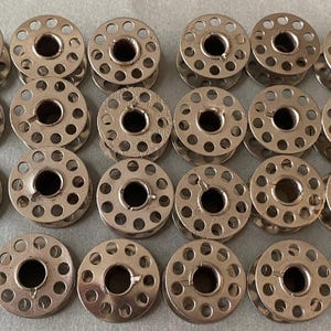 Pode incluir: Uma coleção de bobinas de máquina de costura em metal prateado. Cada bobina tem um orifício central e vários orifícios menores ao redor. As bobinas estão dispostas em fileiras e colunas.