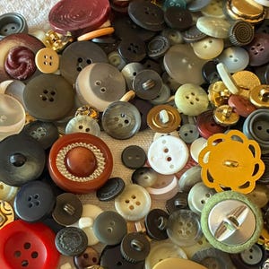 Puede incluir: Una gran colección de botones variados de diferentes formas, tamaños y colores, incluyendo rojo, amarillo, marrón, negro, blanco y verde. Los botones presentan distintas texturas, acabados y números de agujeros, desde discos planos hasta diseños ornamentados.