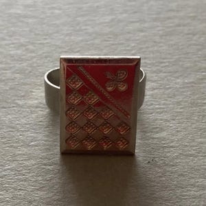 May include: Silver ring with a rectangular face. The face has a red background, a diagonal line, and a four-leaf clover design. Below the line are checkered squares.