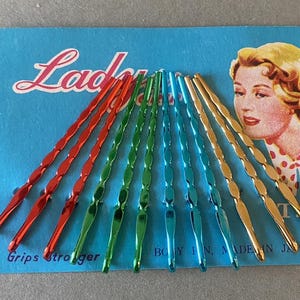 Pode incluir: Um cartão vintage de grampos de cabelo em vermelho, verde, azul e dourado. O cartão é azul claro com a palavra "Ladies" em escrita e uma ilustração retrô de uma mulher. O texto "Grips stronger" também é visível.
