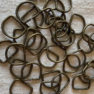 Könnte beinhalten: Eine Ansammlung von D-Ringen aus bronzefarbenem Metall, die in Form von rund bis quadratisch variieren. Die Ringe sind verstreut und zeigen ihr offenes Design, ideal zum Befestigen von Riemen oder anderen Beschlägen. Das Metall hat ein leicht gealtertes Aussehen.