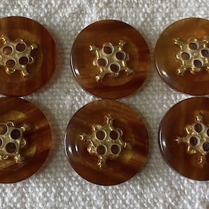 Könnte beinhalten: Sechs runde, braune Knöpfe mit einem goldfarbenen, verzierten Mitteldesign. Die Knöpfe haben eine glänzende Oberfläche und sind in einem ordentlichen Muster angeordnet, geeignet zum Nähen oder Basteln.